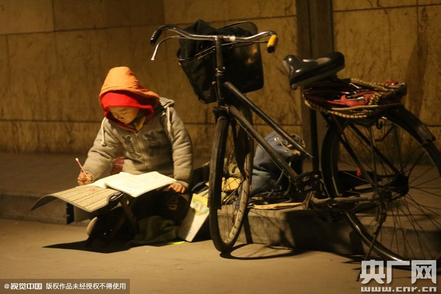 鄭州中原路京廣鐵路大橋下涵洞的馬路牙子邊，一位戴著紅色辣椒帽的小男孩趴在一個小馬扎上，上面鋪著一張車墊，在微弱的燈光下寫作業(yè)