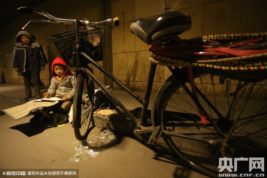 2015年11月8日晚，鄭州，7歲男孩涵洞橋下陪父母練攤，路邊燈下坐小馬扎上寫作業(yè)。